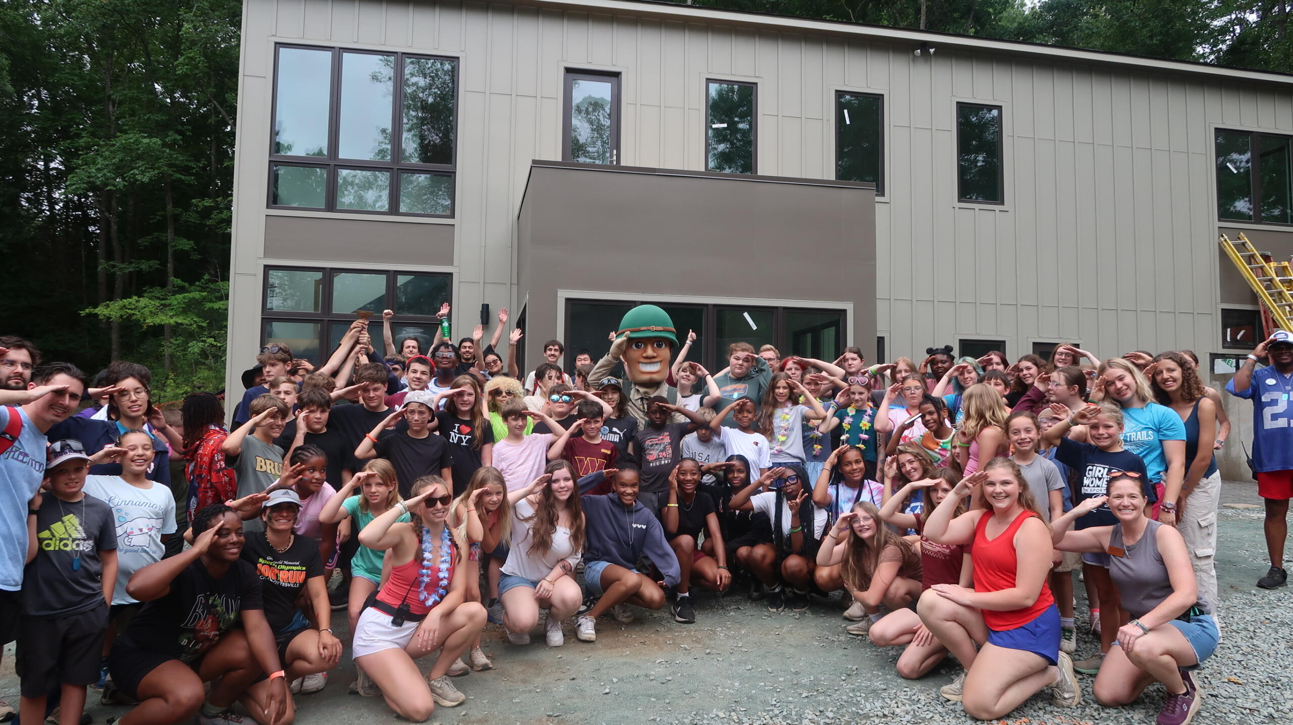 Large group of children posing outdoors in front of a modern building, with a mascot in a helmet at center, cheering and smiling.