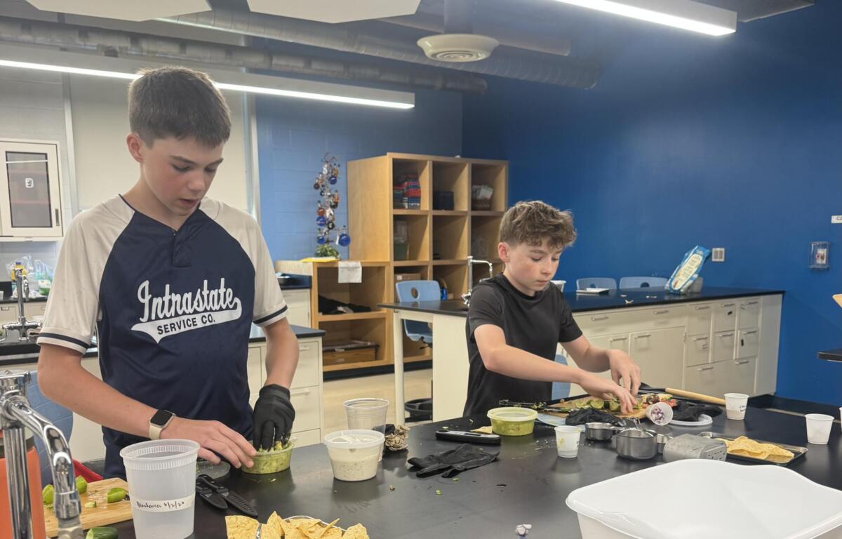 Two students in a classroom kitchen prep area chop vegetables at a dark countertop with bowls and chips nearby.