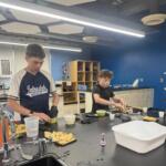 Two students in a classroom kitchen prep area chop vegetables at a dark countertop with bowls and chips nearby.