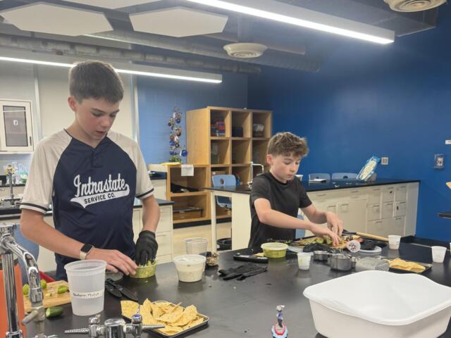 Two students in a classroom kitchen prep area chop vegetables at a dark countertop with bowls and chips nearby.