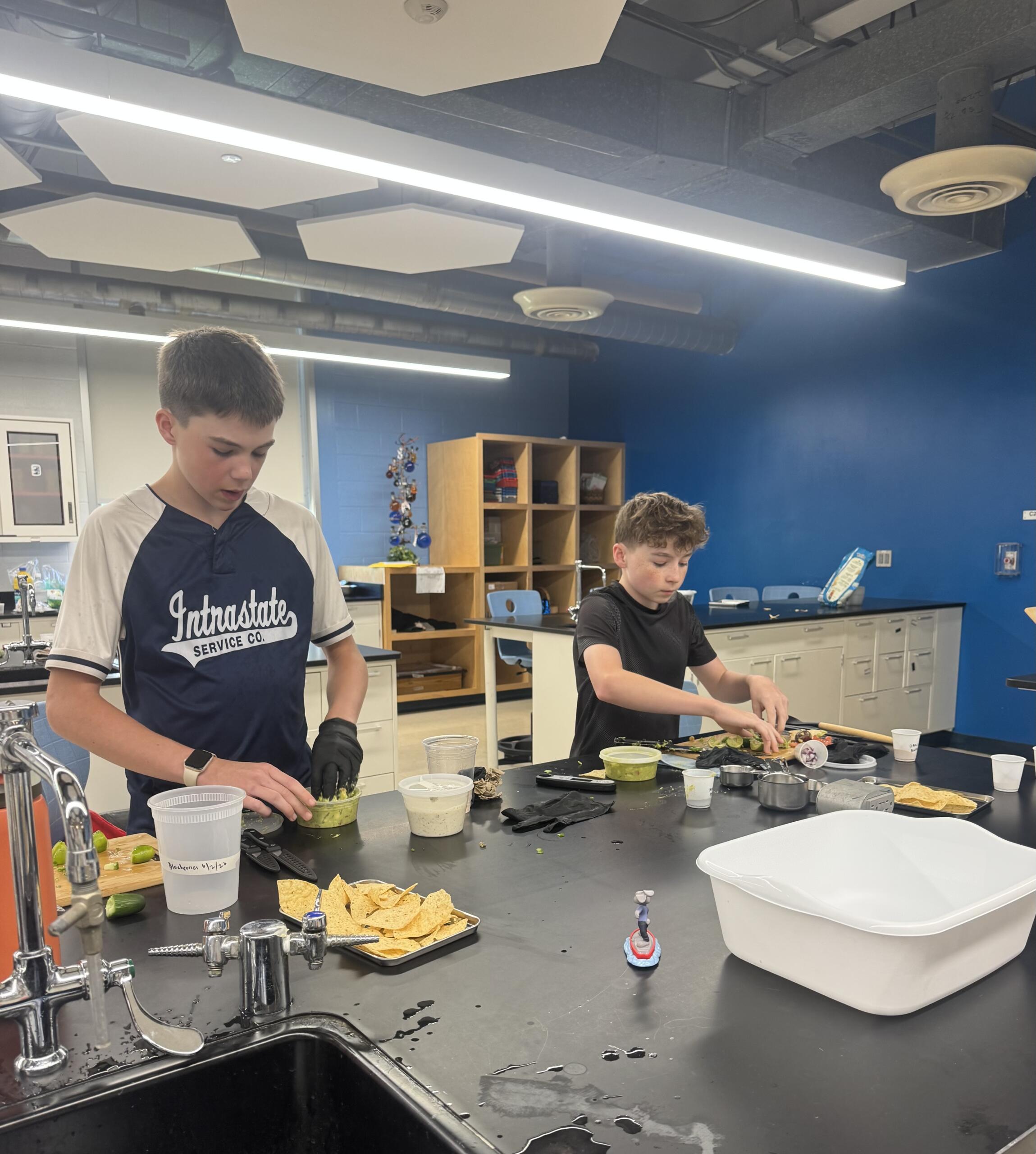 Two students in a classroom kitchen prep area chop vegetables at a dark countertop with bowls and chips nearby.