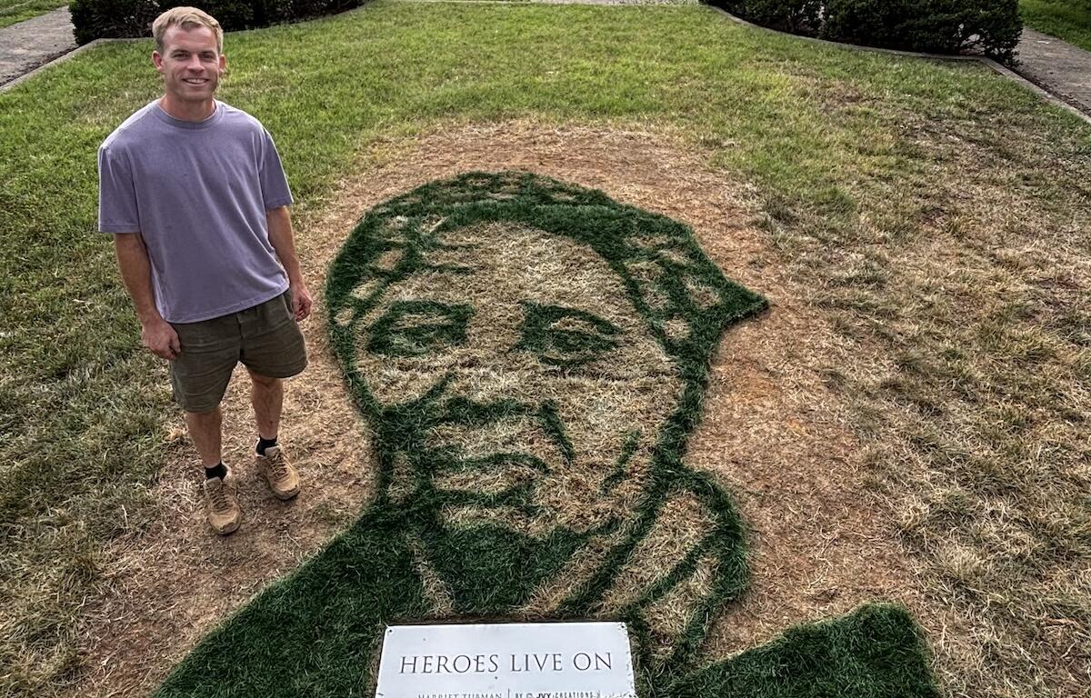 Man in a lavender shirt and shorts stands on a lawn beside a large grass portrait of a man's head, with a plaque reading 'HEROES LIVE ON' in front.