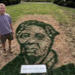 Man in a lavender shirt and shorts stands on a lawn beside a large grass portrait of a man's head, with a plaque reading 'HEROES LIVE ON' in front.