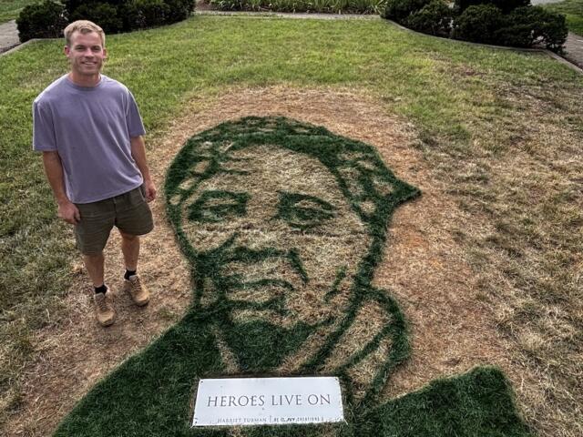 Man in a lavender shirt and shorts stands on a lawn beside a large grass portrait of a man's head, with a plaque reading 'HEROES LIVE ON' in front.