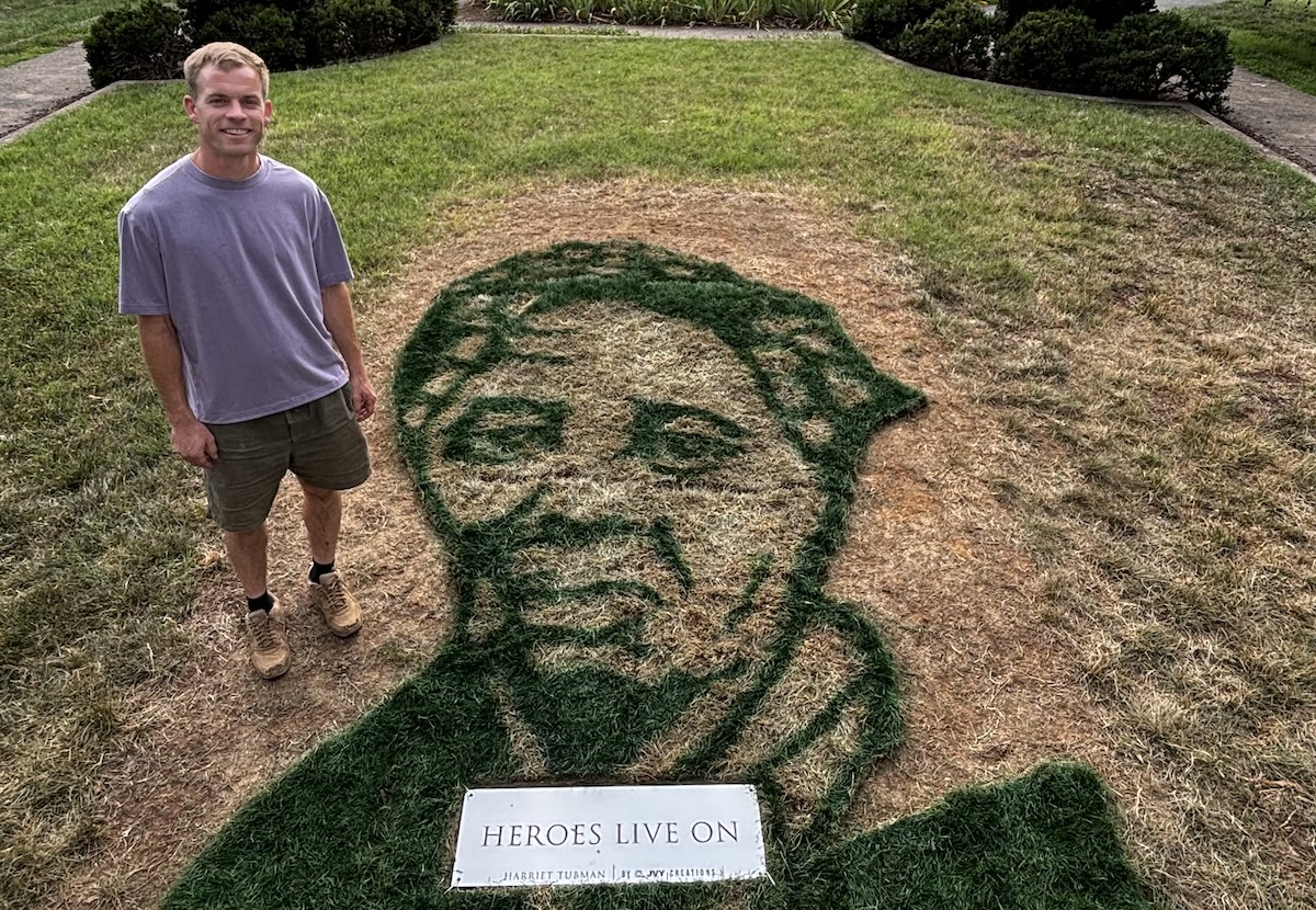 Man in a lavender shirt and shorts stands on a lawn beside a large grass portrait of a man's head, with a plaque reading 'HEROES LIVE ON' in front.