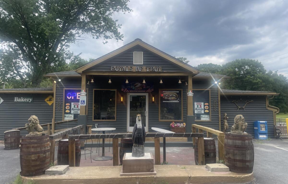 Exterior of Royals of Troy with an open sign, rustic outdoor seating, and wooden barrels under a cloudy sky.