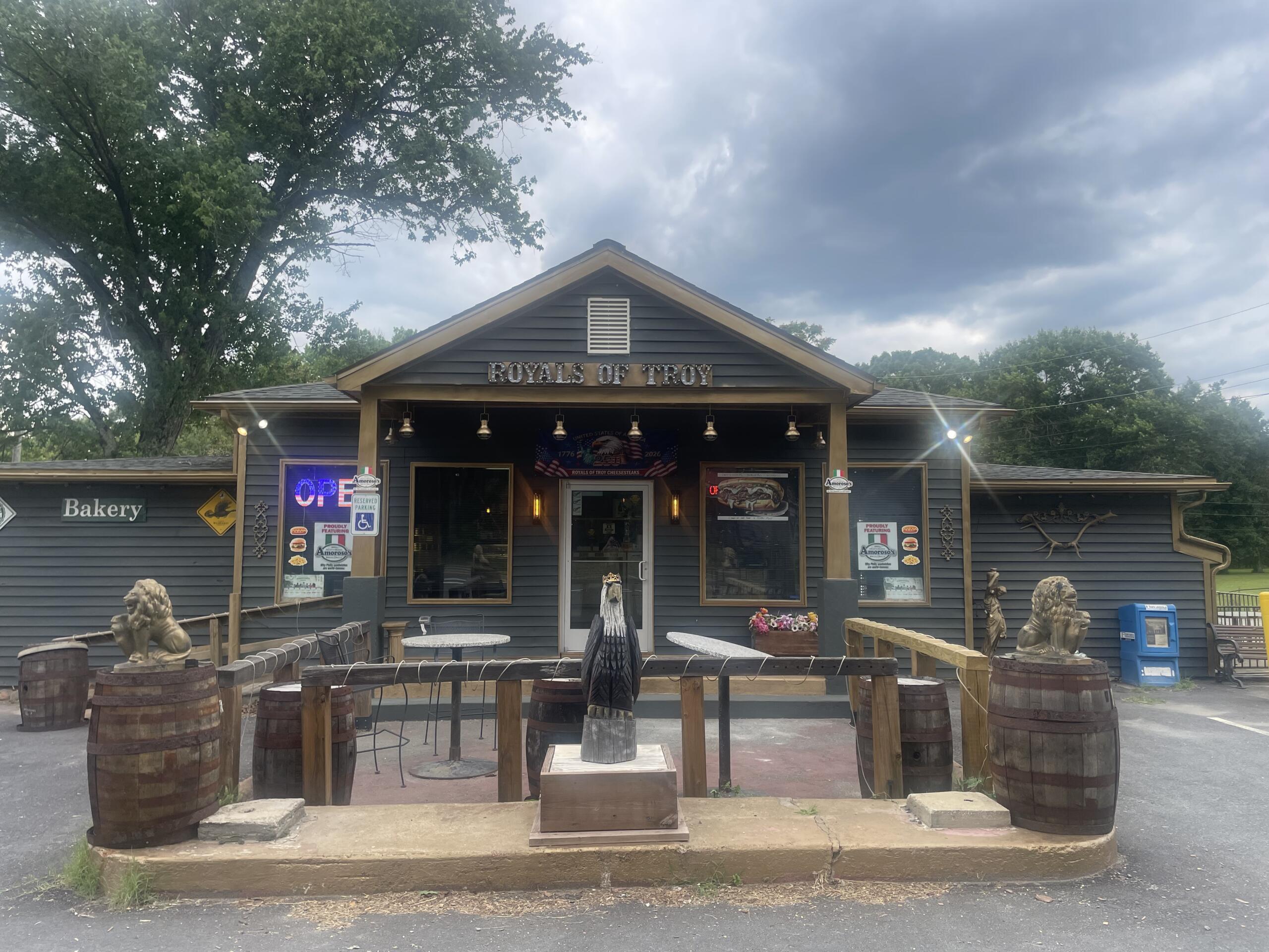 Exterior of Royals of Troy with an open sign, rustic outdoor seating, and wooden barrels under a cloudy sky.