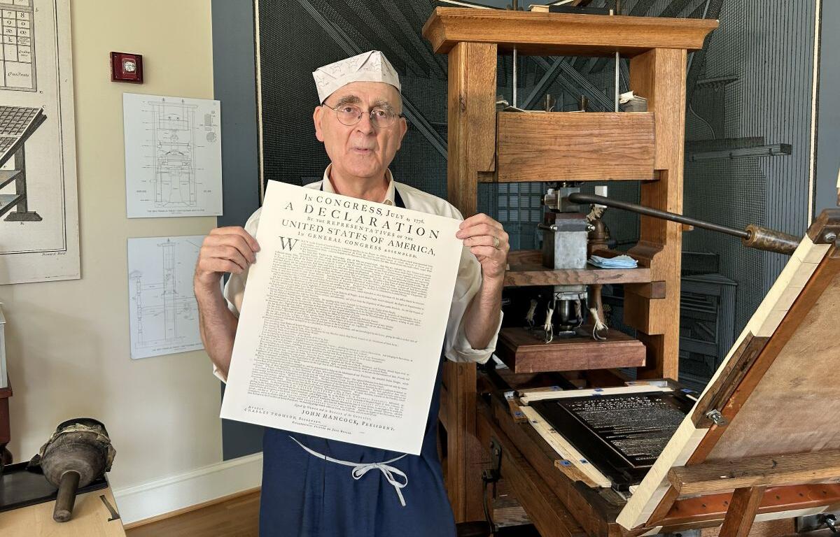 Older man in a white paper hat and apron holds a large printed Declaration beside a vintage printing press in a workshop setting.