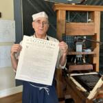 Older man in a white paper hat and apron holds a large printed Declaration beside a vintage printing press in a workshop setting.