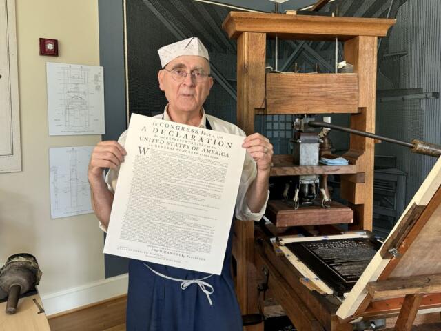 Older man in a white paper hat and apron holds a large printed Declaration beside a vintage printing press in a workshop setting.