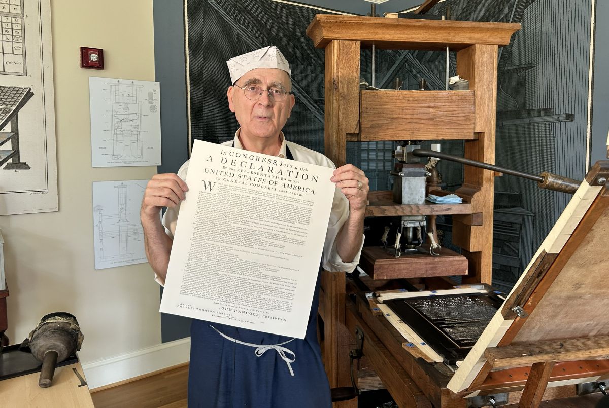 Older man in a white paper hat and apron holds a large printed Declaration beside a vintage printing press in a workshop setting.
