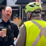 Policeman in tactical vest holding a coffee cup speaks with a person wearing a bright safety vest and helmet outdoors.