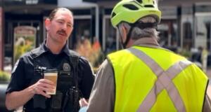 Policeman in tactical vest holding a coffee cup speaks with a person wearing a bright safety vest and helmet outdoors.