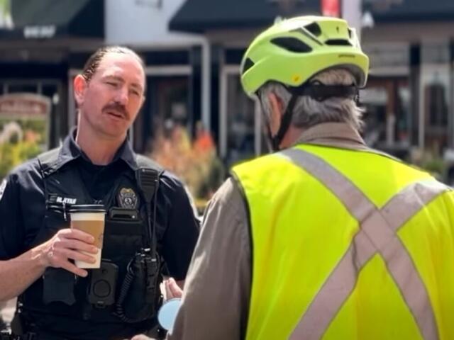 Policeman in tactical vest holding a coffee cup speaks with a person wearing a bright safety vest and helmet outdoors.