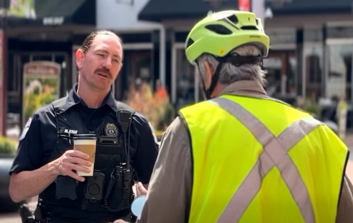 Policeman in tactical vest holding a coffee cup speaks with a person wearing a bright safety vest and helmet outdoors.