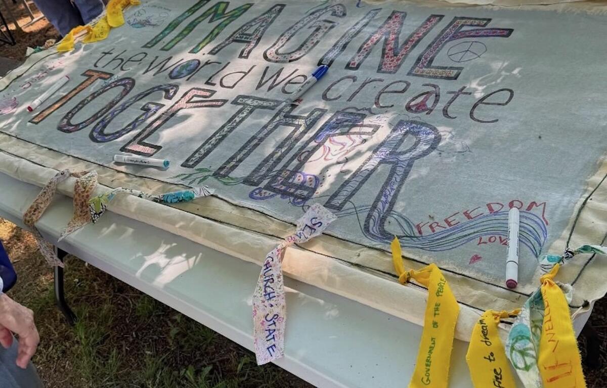 Handmade banner laid over a folding table with colorful, large lettering promoting a message about peace, freedom, and imagining a better world; markers and fabric scraps are nearby.