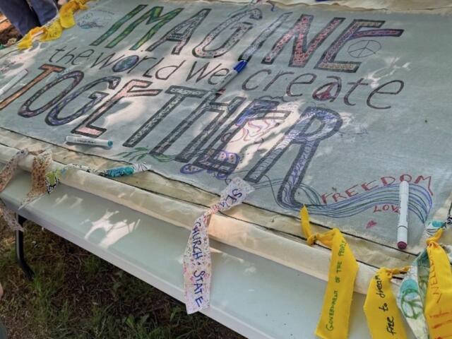 Handmade banner laid over a folding table with colorful, large lettering promoting a message about peace, freedom, and imagining a better world; markers and fabric scraps are nearby.
