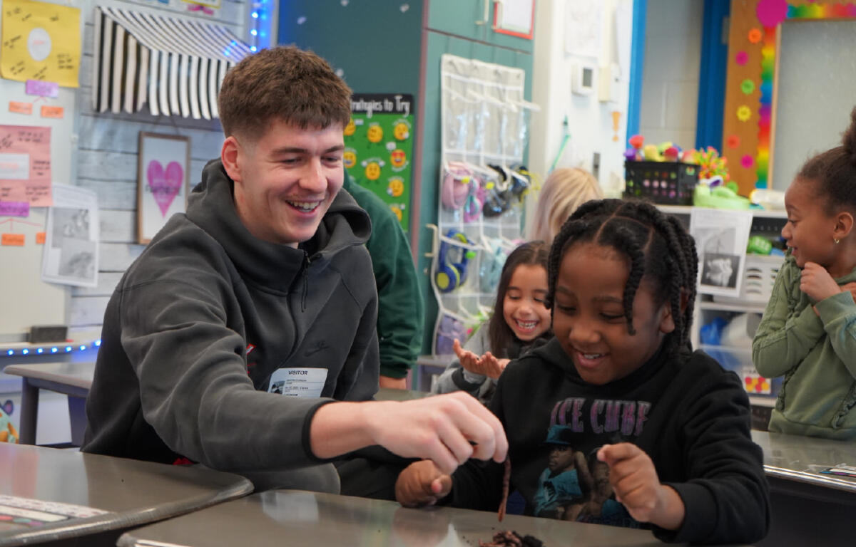 Smiling man and two girls at a desk share a hands-on activity in a classroom; they pass a brown object to each other while other students watch happily.