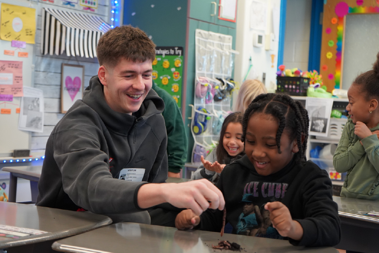 Smiling man and two girls at a desk share a hands-on activity in a classroom; they pass a brown object to each other while other students watch happily.