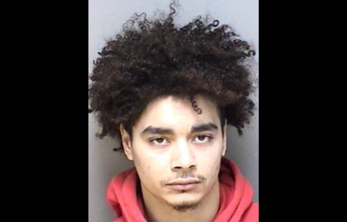 Mugshot of a young man with thick curly hair wearing a red hoodie, looking straight at the camera with a neutral expression.
