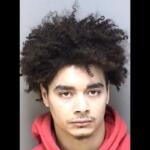 Mugshot of a young man with thick curly hair wearing a red hoodie, looking straight at the camera with a neutral expression.