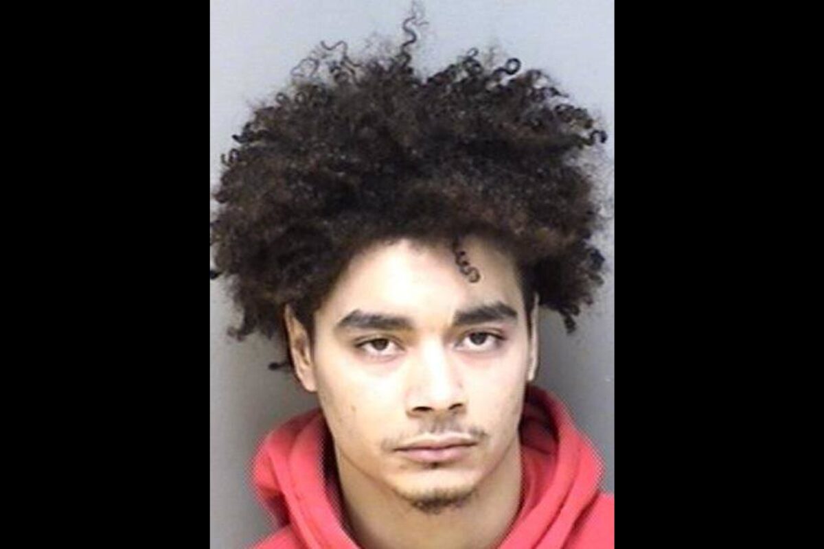 Mugshot of a young man with thick curly hair wearing a red hoodie, looking straight at the camera with a neutral expression.