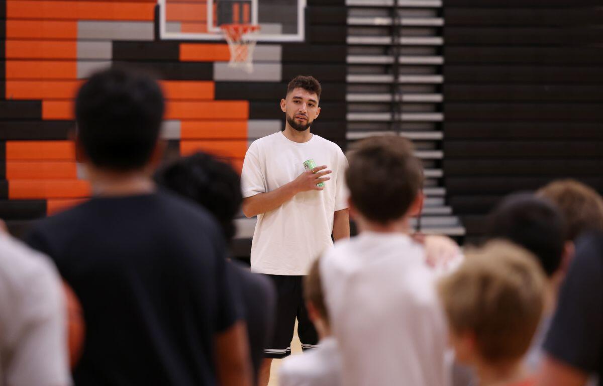 Coach in a white t-shirt speaking to a group in a gym, holding a drink, with a basketball hoop in the background.