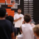 Coach in a white t-shirt speaking to a group in a gym, holding a drink, with a basketball hoop in the background.