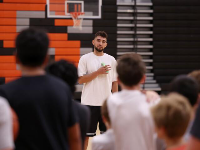 Coach in a white t-shirt speaking to a group in a gym, holding a drink, with a basketball hoop in the background.