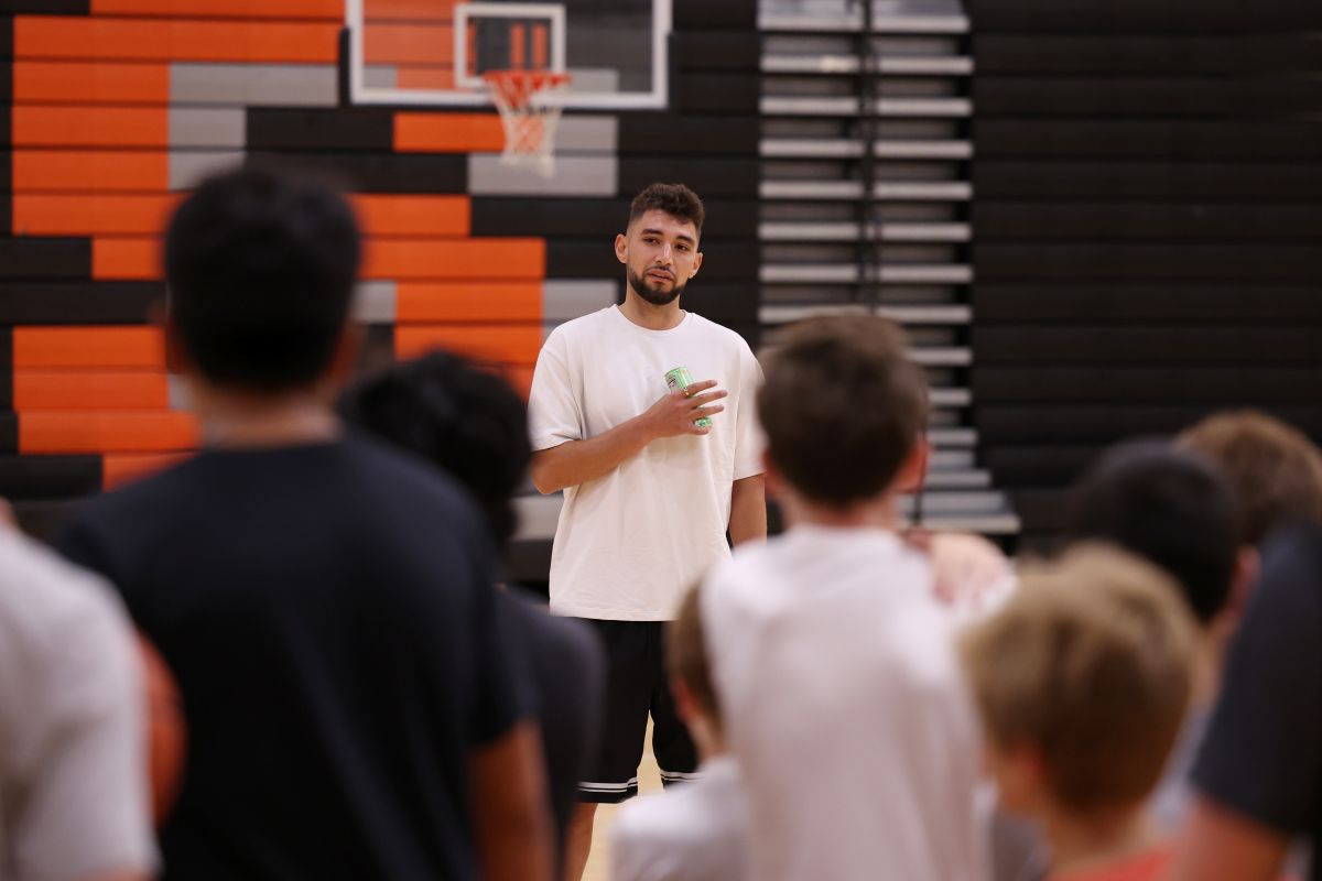 Coach in a white t-shirt speaking to a group in a gym, holding a drink, with a basketball hoop in the background.