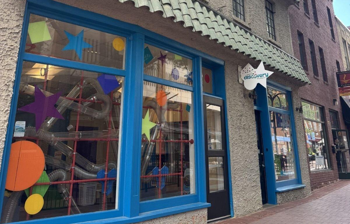 Front of the Discovery Museum with bright blue window frames and colorful star shapes on the glass; ducts and tubes visible inside.ans