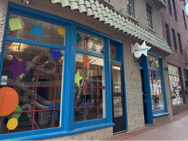 Front of the Discovery Museum with bright blue window frames and colorful star shapes on the glass; ducts and tubes visible inside.ans
