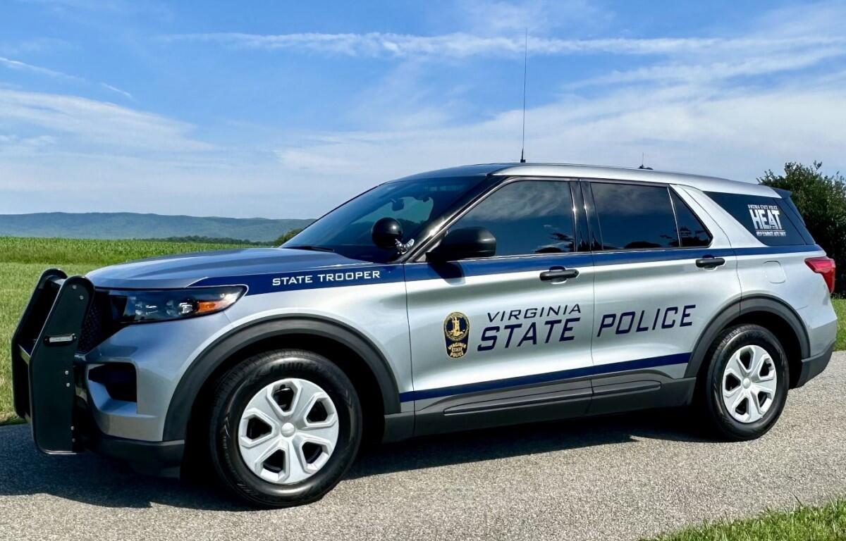 Virginia State Police SUV parked on a rural road with a blue sky and green fields in the background, showing 'STATE TROOPER' lettering on the side.