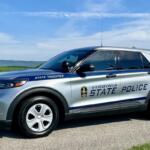 Virginia State Police SUV parked on a rural road with a blue sky and green fields in the background, showing 'STATE TROOPER' lettering on the side.