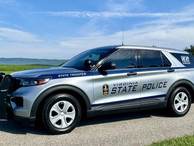 Virginia State Police SUV parked on a rural road with a blue sky and green fields in the background, showing 'STATE TROOPER' lettering on the side.