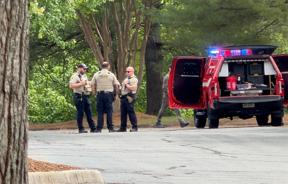 Police officers in beige vests speak near a red emergency vehicle with flashing lights in a wooded area.