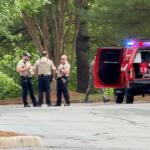 Police officers in beige vests speak near a red emergency vehicle with flashing lights in a wooded area.