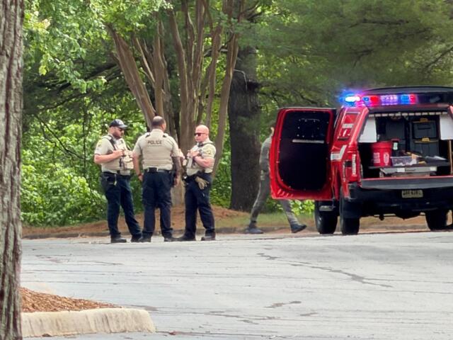 Police officers in beige vests speak near a red emergency vehicle with flashing lights in a wooded area.