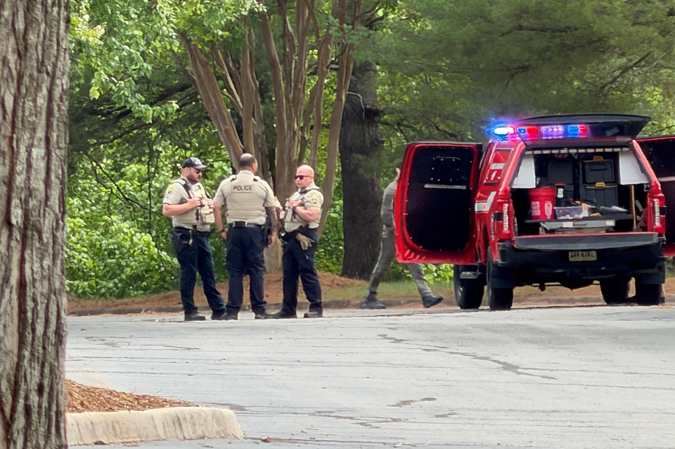 Police officers in beige vests speak near a red emergency vehicle with flashing lights in a wooded area.