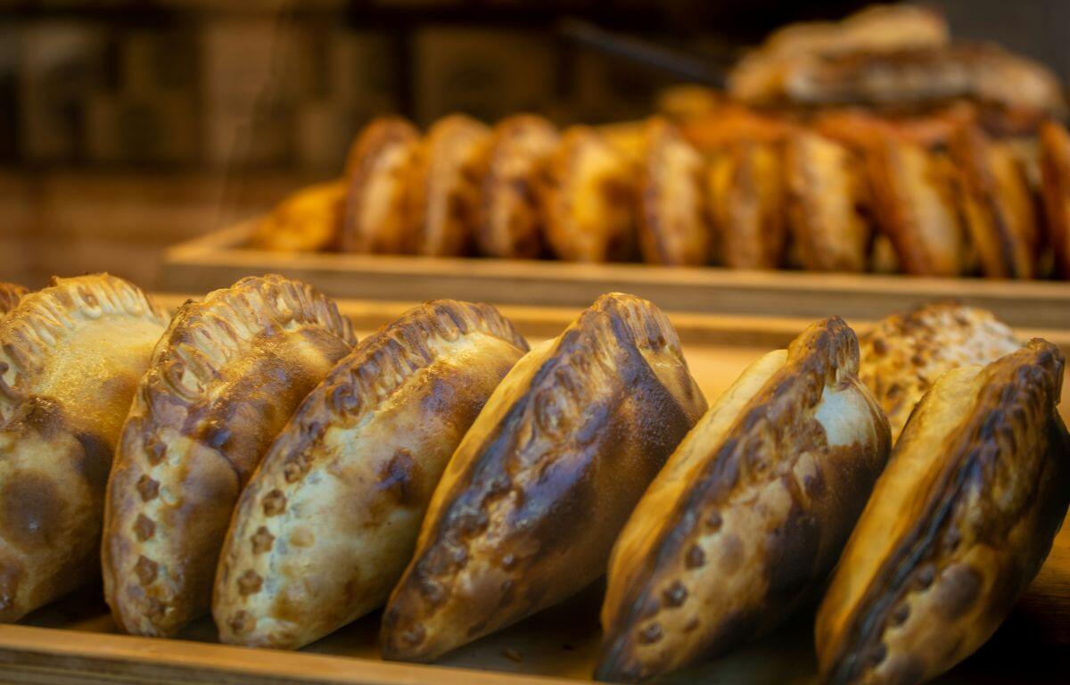 Row of golden-brown empanadas or turnovers on a wooden tray in a warmly lit bakery display, with more pastries blurred in the background