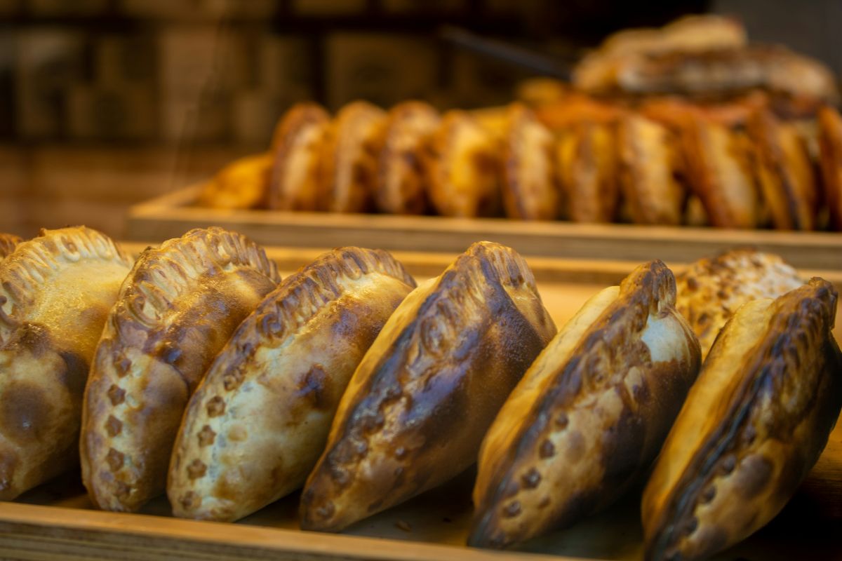 Row of golden-brown empanadas or turnovers on a wooden tray in a warmly lit bakery display, with more pastries blurred in the background