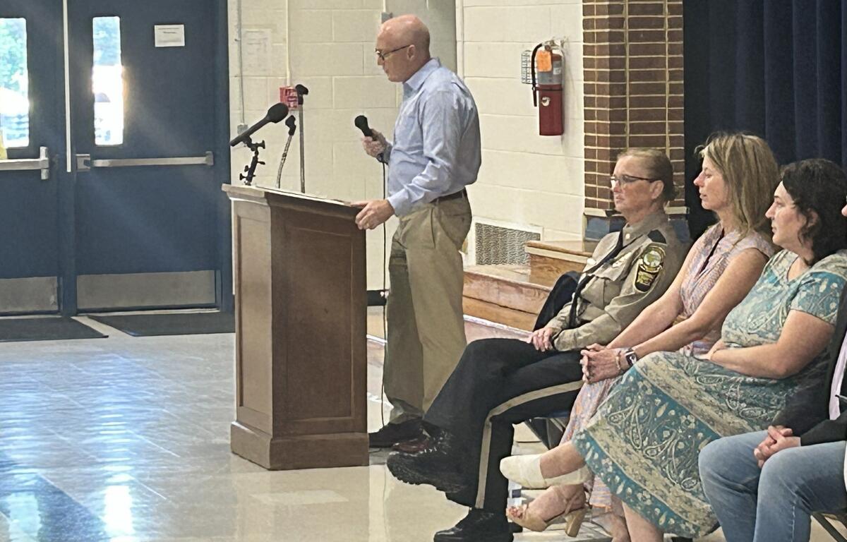 A man stands at a wooden lectern with two microphones, speaking to an audience in a school gym or hall, while a uniformed officer and three women sit nearby.