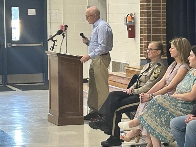 A man stands at a wooden lectern with two microphones, speaking to an audience in a school gym or hall, while a uniformed officer and three women sit nearby.