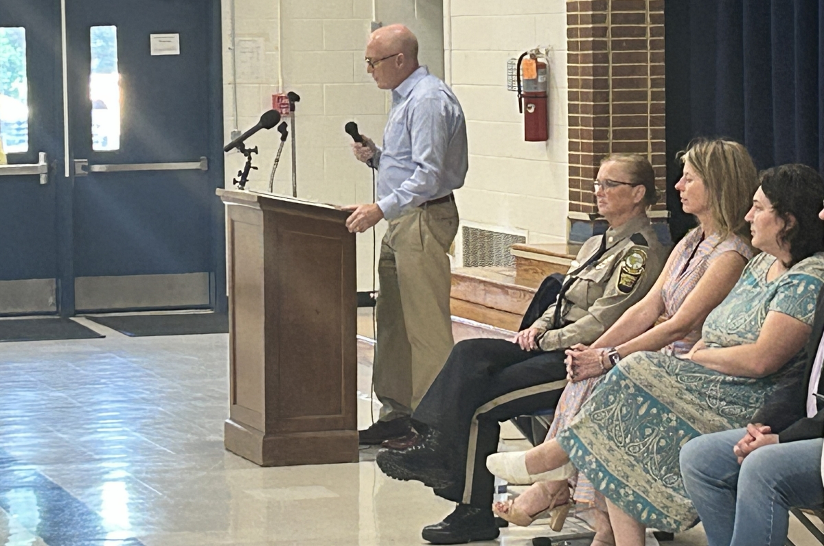 A man stands at a wooden lectern with two microphones, speaking to an audience in a school gym or hall, while a uniformed officer and three women sit nearby.