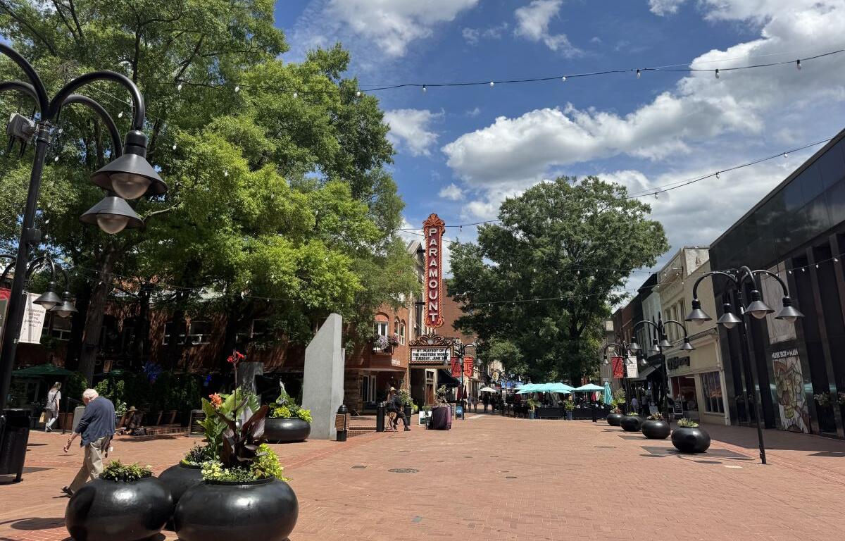 Downtown pedestrian street with a tall Paramount theater sign, string lights, and outdoor seating under umbrellas, daytime.