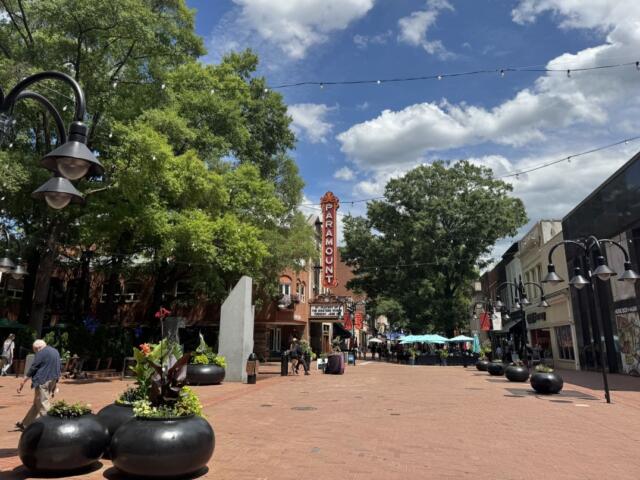 Downtown pedestrian street with a tall Paramount theater sign, string lights, and outdoor seating under umbrellas, daytime.