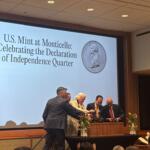 Group of adults at a podium during a ceremony, large screen behind them reads 'U.S. Mint at Monticello: Celebrating the Declaration of Independence Quarter' with a coin image.