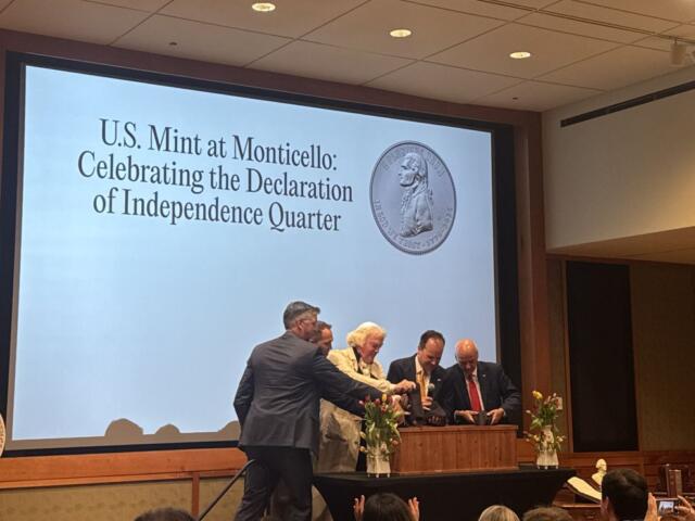 Group of adults at a podium during a ceremony, large screen behind them reads 'U.S. Mint at Monticello: Celebrating the Declaration of Independence Quarter' with a coin image.