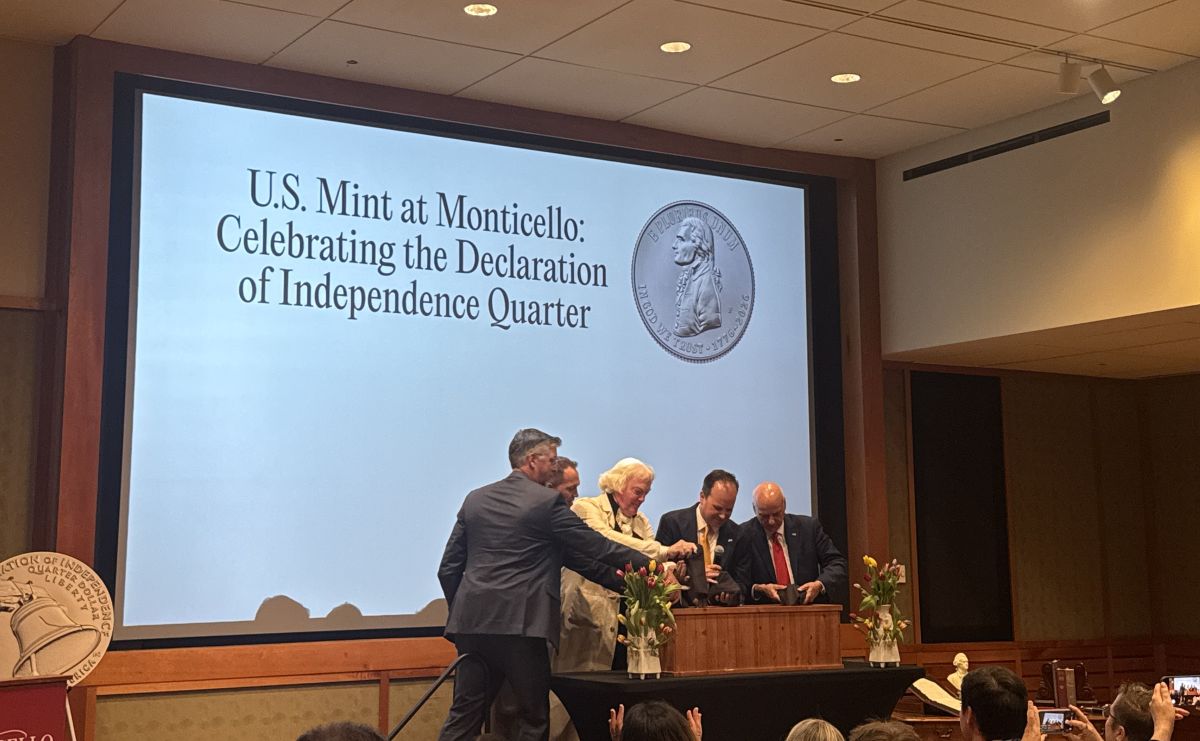 Group of adults at a podium during a ceremony, large screen behind them reads 'U.S. Mint at Monticello: Celebrating the Declaration of Independence Quarter' with a coin image.