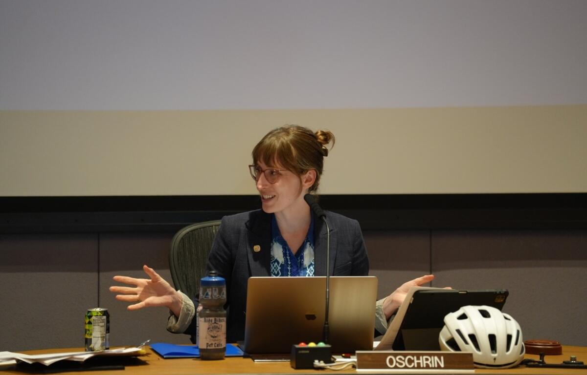 Woman in a blazer speaks at a panel desk with a laptop and microphone, bike helmet on the desk nearby, OSCHRIN nameplate visible.